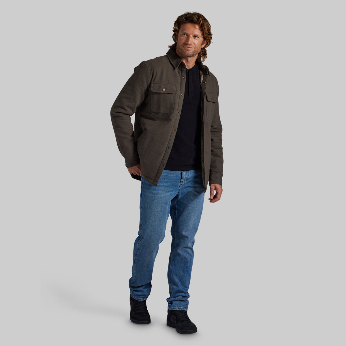 A man modeling a dark olive green utility jacket over a black shirt, blue jeans, and black boots, standing against a solid light gray background.