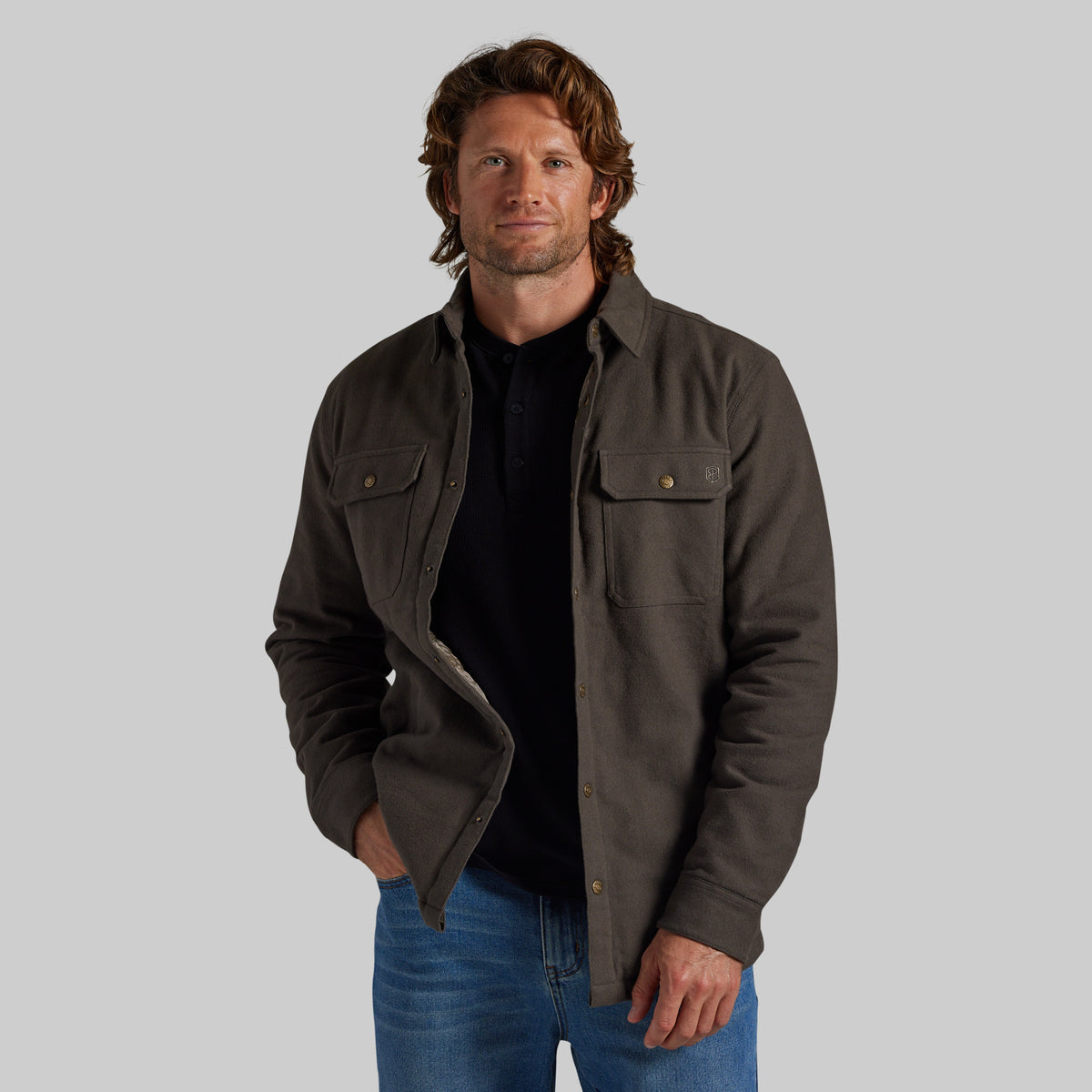 A man wearing a dark brown button-up overshirt jacket over a black shirt and blue jeans, standing against a solid gray background.
