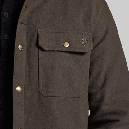 Close-up of a dark olive brown textured overshirt jacket with brass buttons and a small embroidered shield logo on the chest pocket, worn over a dark shirt.