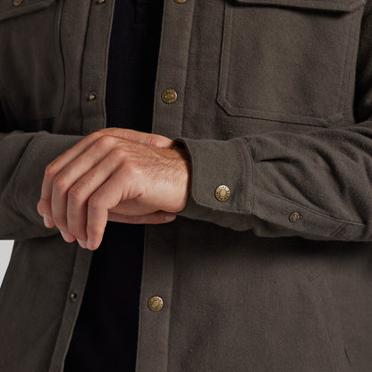 Close-up of a person wearing a dark olive-brown overshirt with brass buttons over a black shirt, showing the hands adjusting or fastening the cuff.