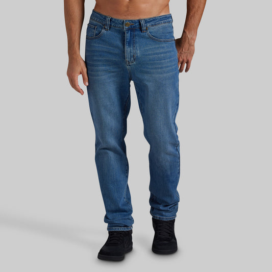 A person modeling a pair of medium blue slim-fit jeans with black sneakers against a solid light gray studio background.