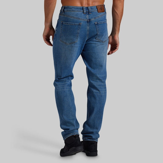 Rear view of a man wearing blue slim-fit jeans with black boots, shown against a solid gray studio background.
