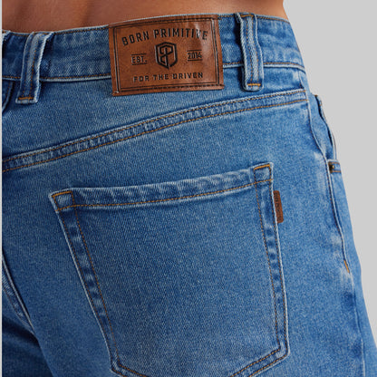 Close-up of the back waistband and pocket area of blue denim jeans on a model, showing a brown leather patch with the Born Primitive logo.
