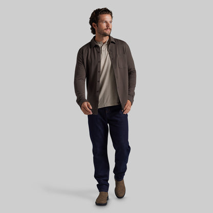 A man modeling a dark brown overshirt over a beige shirt, paired with navy blue pants and brown boots, against a solid gray studio background.