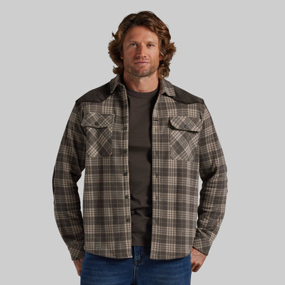 A man wearing a brown and beige plaid button-up shirt jacket with dark brown shoulder panels, layered over a dark t-shirt and blue jeans, against a solid gray background.