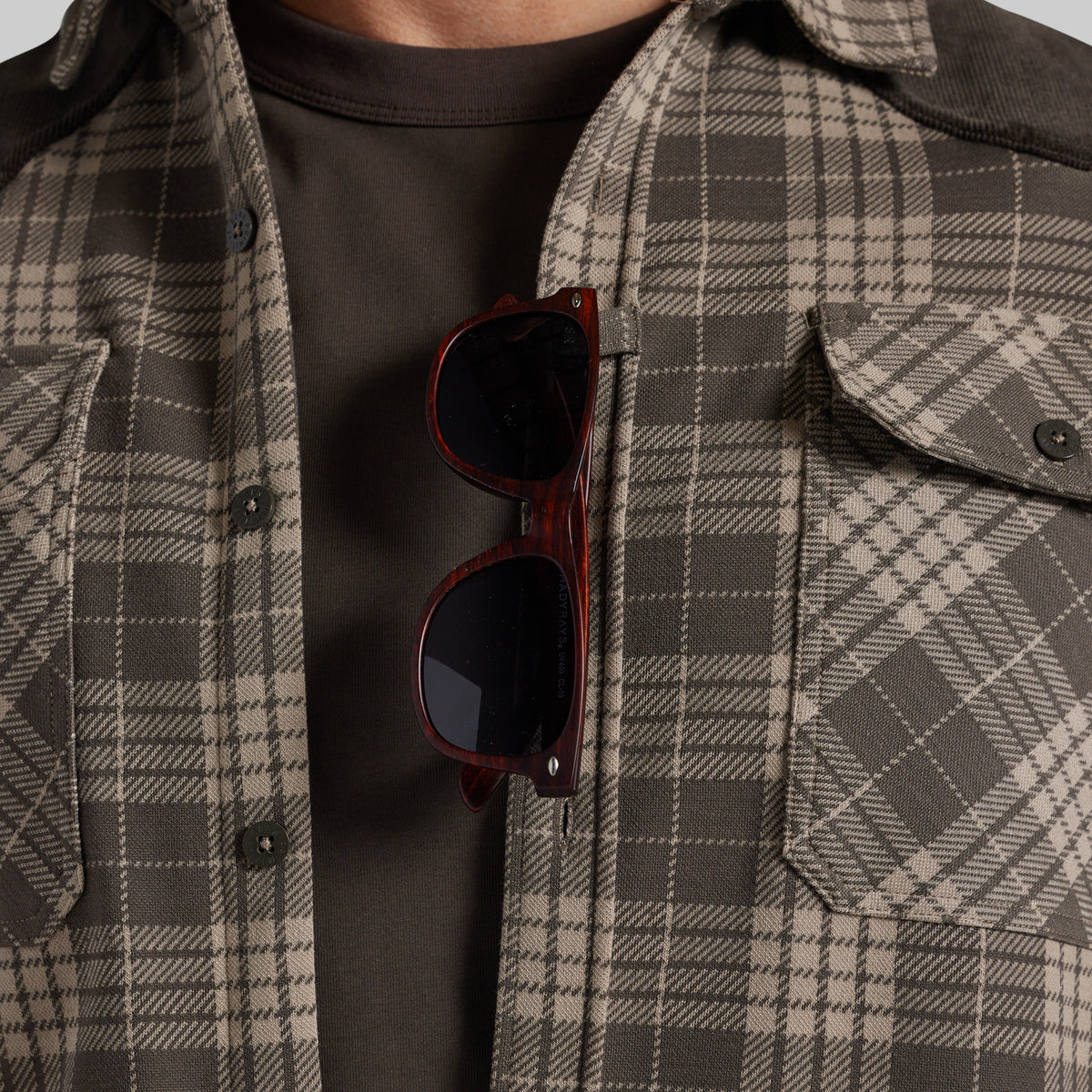 A close-up of a person wearing a brown and beige plaid overshirt over a dark brown t-shirt, with tortoiseshell sunglasses hanging from the placket.