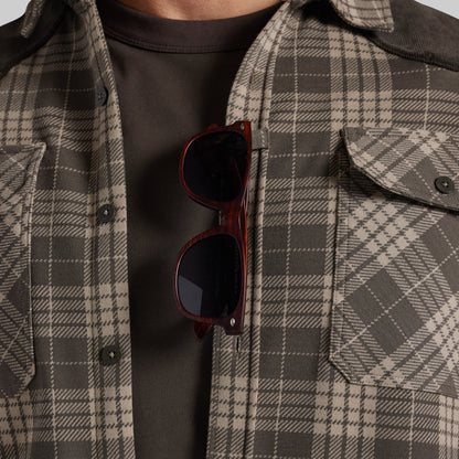 A close-up of a person wearing a brown and beige plaid overshirt over a dark brown t-shirt, with tortoiseshell sunglasses hanging from the placket.