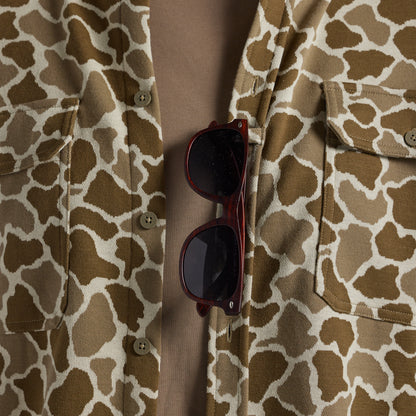 A close-up of a person wearing a brown and beige frog camo button-up shirt, with dark sunglasses tucked into the open front pocket area.