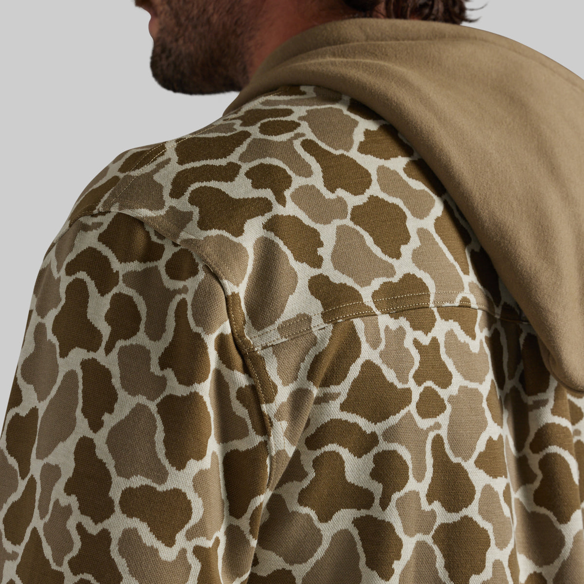 Close-up of a person wearing a brown and beige frog camo patterned hoodie with a solid brown hood, shown from the back and side against a light gray background.