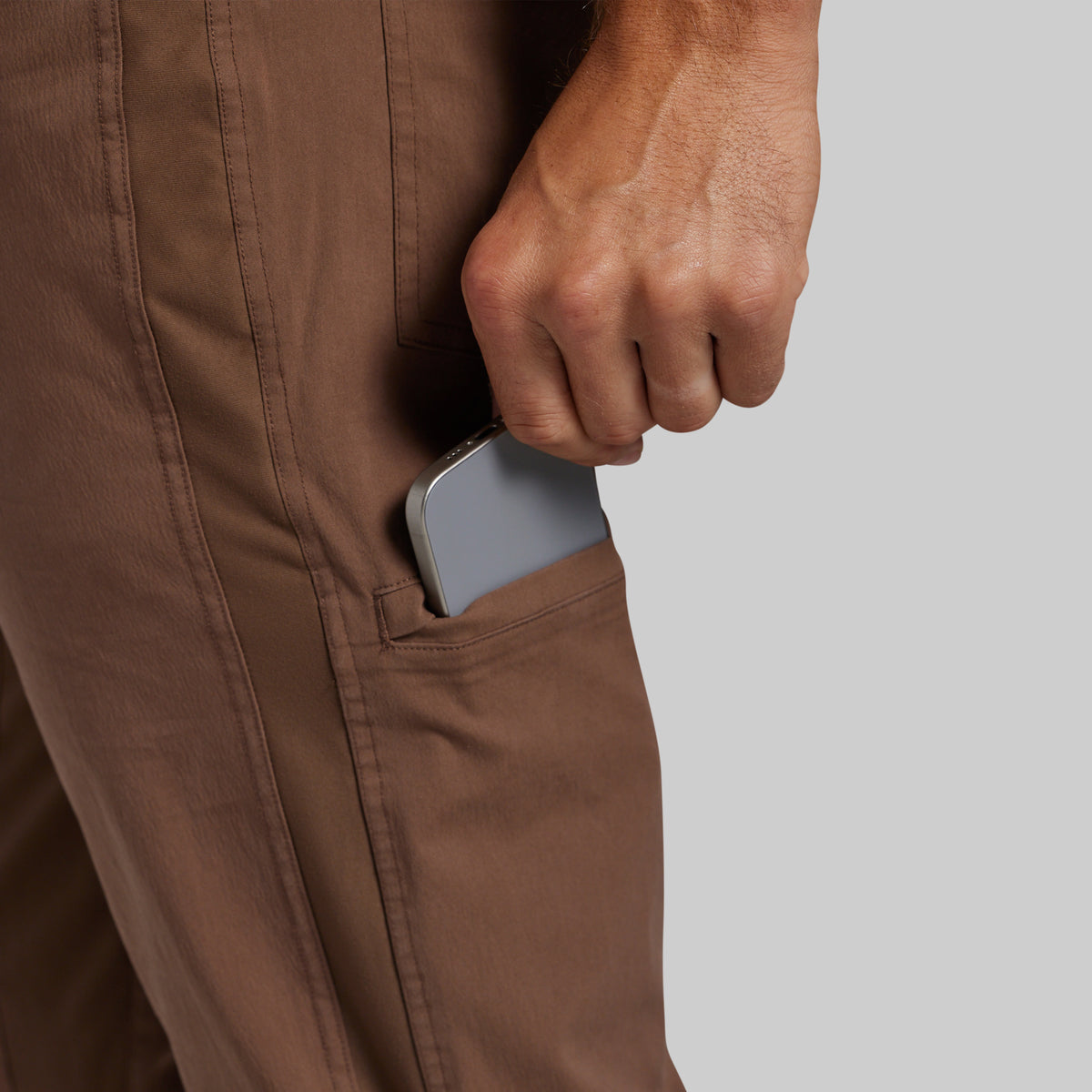 Close-up of a person wearing brown pants, pulling a silver smartphone out of a side pocket against a light gray background.