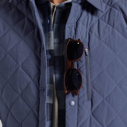 Close-up of a navy blue diamond-quilted jacket with a chest zip pocket, worn over a plaid shirt. A pair of brown sunglasses rests in the pocket.
