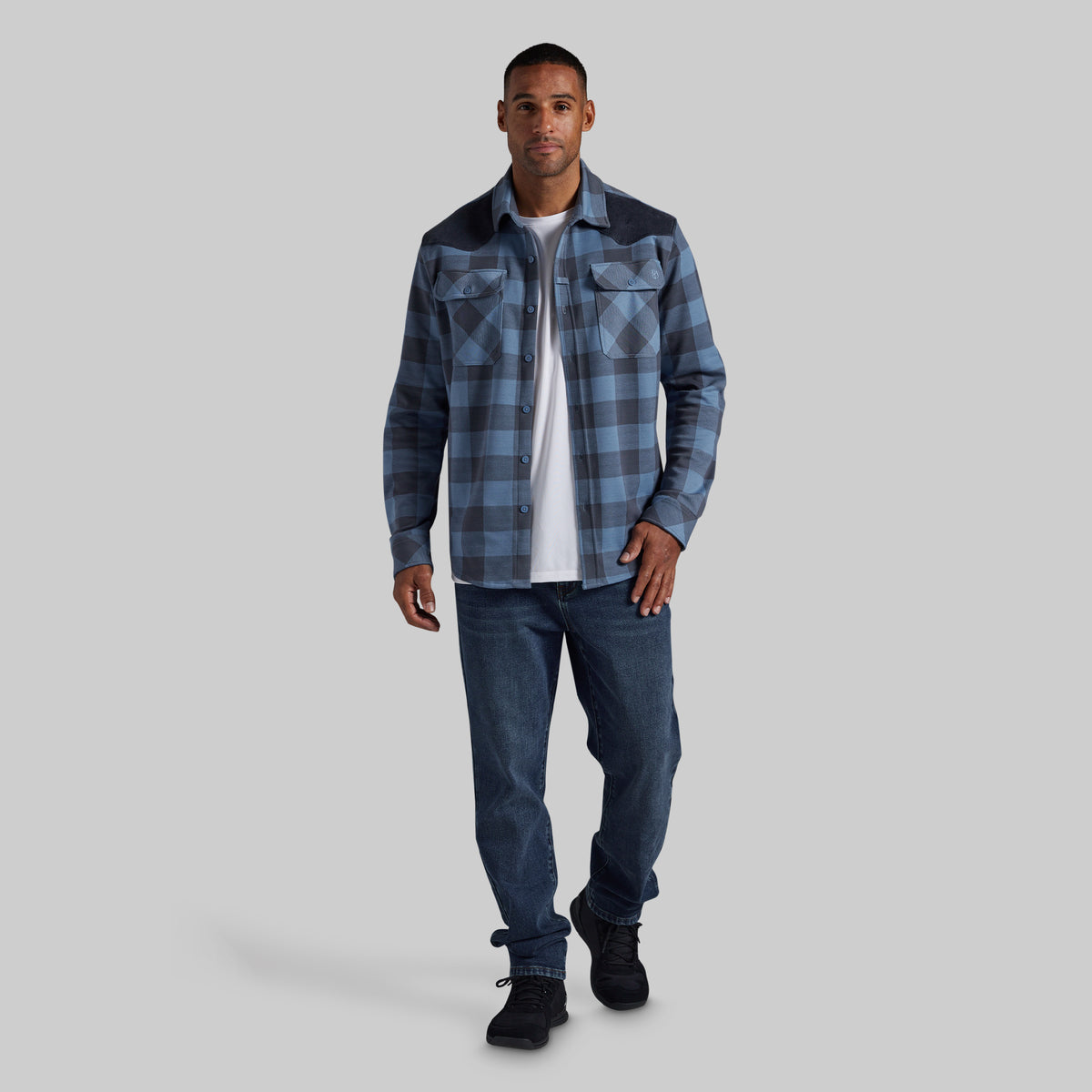 A man modeling a blue and gray plaid flannel shirt with dark navy shoulder panels, worn over a white t-shirt and blue jeans, against a solid gray background.