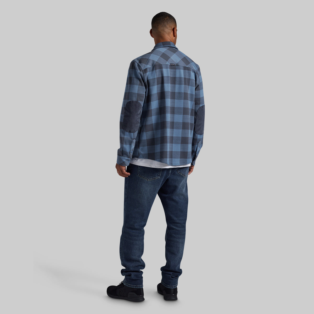 A man viewed from behind wearing a blue and gray plaid button-up shirt with dark blue jeans and black shoes, standing against a solid gray studio background.