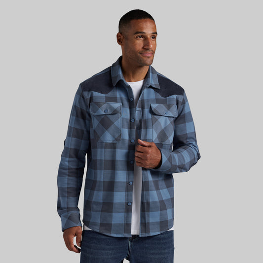 A man wearing a blue and gray plaid button-up shirt with dark navy shoulder panels, layered over a white t-shirt and blue jeans, set against a solid gray background.