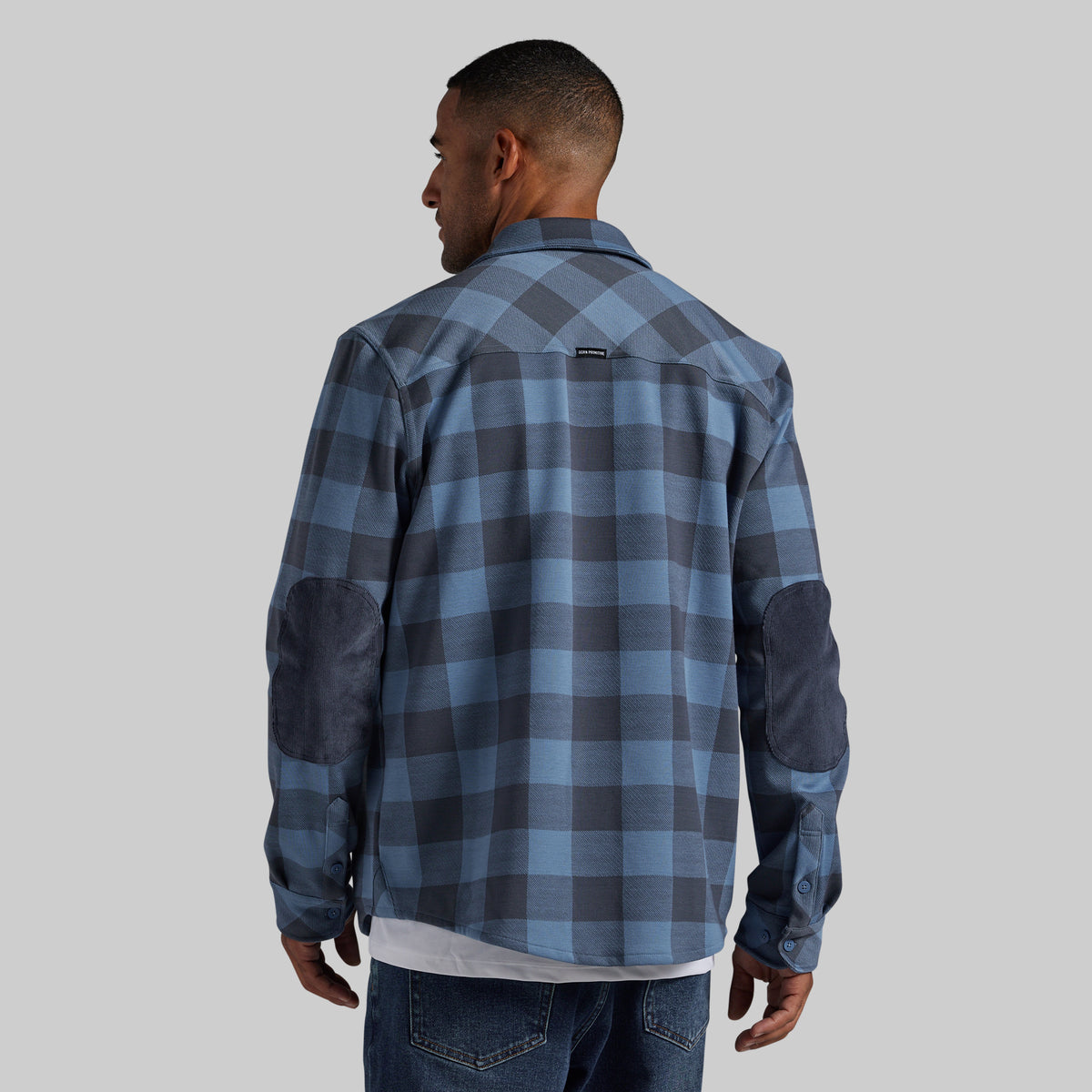 A man viewed from the back wearing a blue and gray plaid flannel shirt with dark blue elbow patches, paired with blue jeans, against a solid gray background.