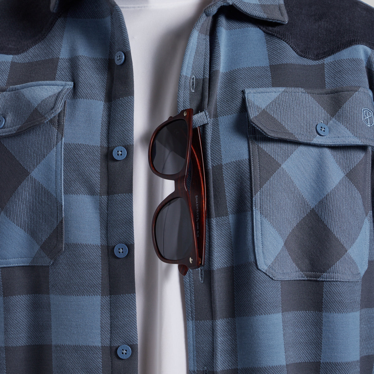 A close-up of a blue and gray plaid button-up shirt with a dark corduroy collar, featuring a chest pocket with a button and a pair of sunglasses hanging from the placket over a white undershirt.