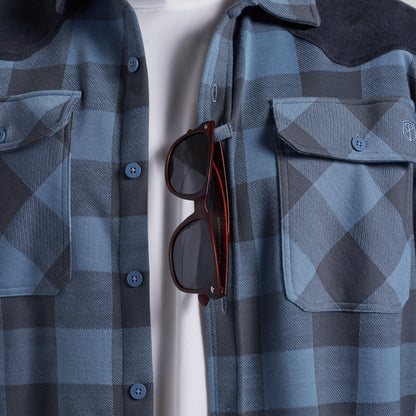 A close-up of a blue and gray plaid button-up shirt with a dark corduroy collar, featuring a chest pocket with a button and a pair of sunglasses hanging from the placket over a white undershirt.
