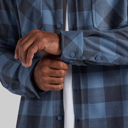 Close-up of a person wearing a blue and gray buffalo plaid flannel shirt, adjusting the cuff of the right sleeve over a white undershirt.