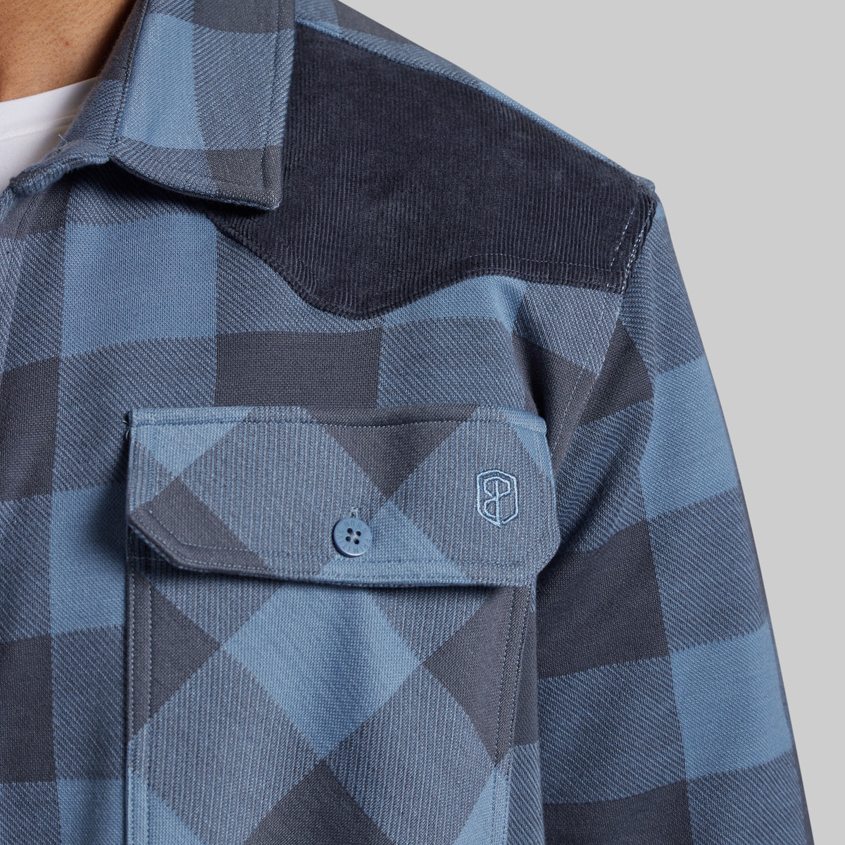 Close-up of a blue and gray plaid western-style shirt jacket with a corduroy yoke and a chest pocket featuring a small embroidered shield logo.
