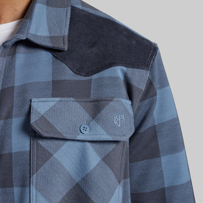 Close-up of a blue and gray plaid western-style shirt jacket with a corduroy yoke and a chest pocket featuring a small embroidered shield logo.
