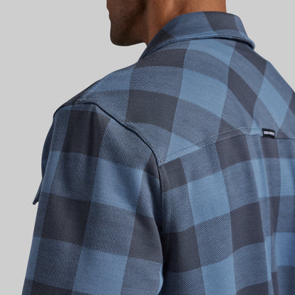Close-up back view of a man wearing a blue and dark gray plaid flannel shirt, showing the fabric texture and a small black brand tag on the upper shoulder.