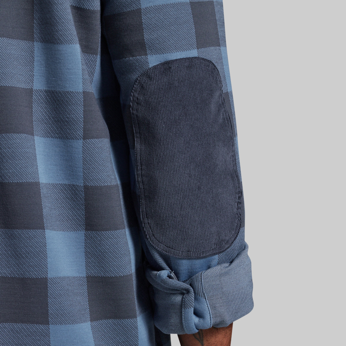Close-up of a blue and gray buffalo plaid flannel shirt with a dark blue corduroy elbow patch and rolled-up cuff, shown on a model against a light gray background.