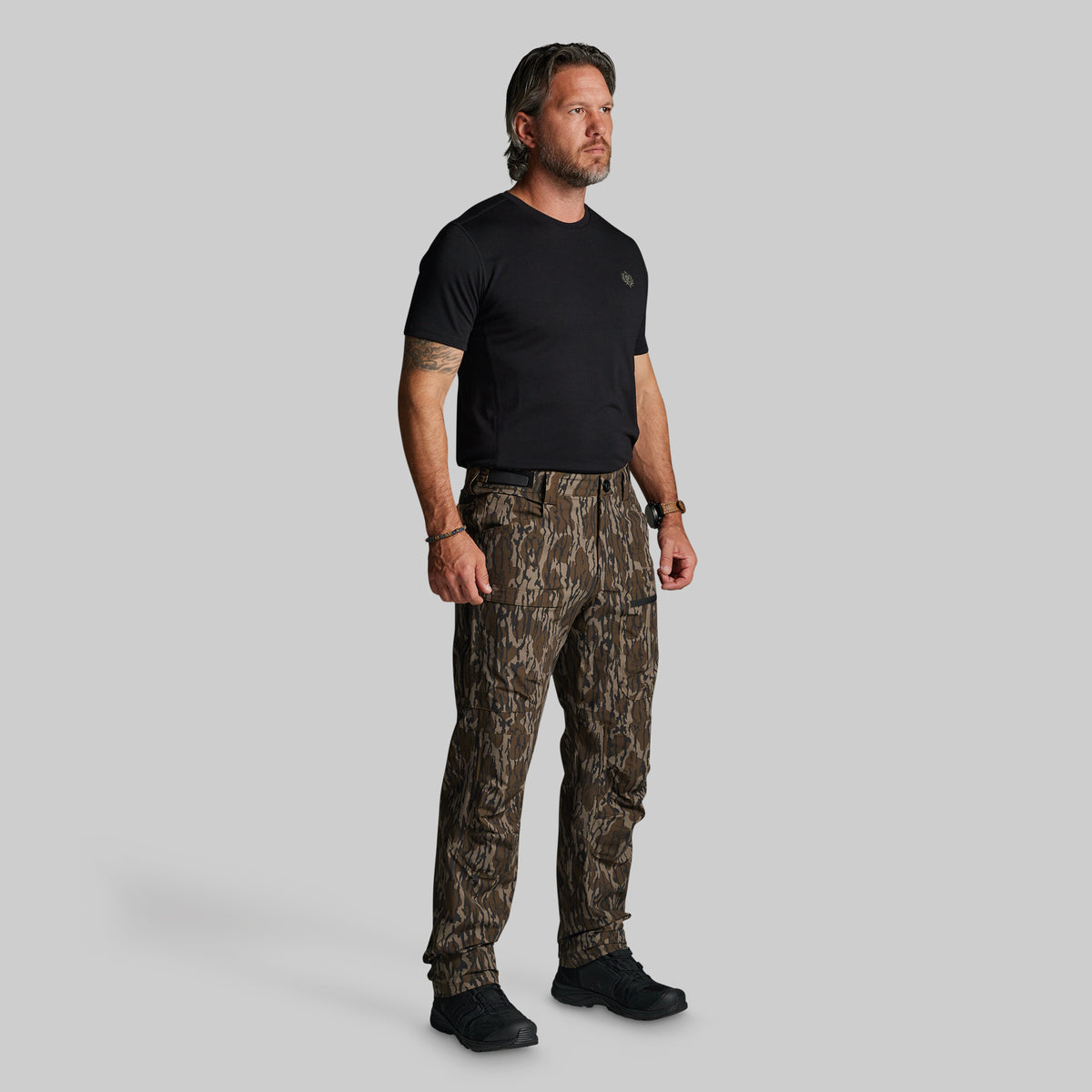 A man modeling a pair of brown and black camouflage pants, paired with a black t-shirt and black shoes, standing against a solid gray background.