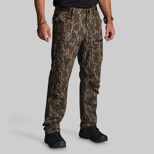 A man wearing camouflage hunting pants with a brown and green pattern, paired with a black shirt and black tactical shoes, shown from the waist down on a solid gray background.