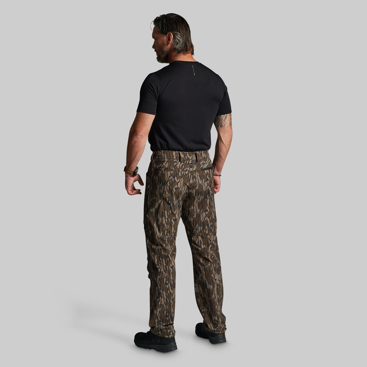 A man viewed from behind wearing camouflage pants, a black t-shirt, and black boots, standing against a solid gray background.