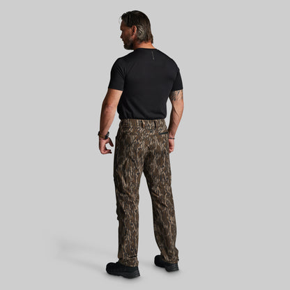A man viewed from behind wearing camouflage pants, a black t-shirt, and black boots, standing against a solid gray background.