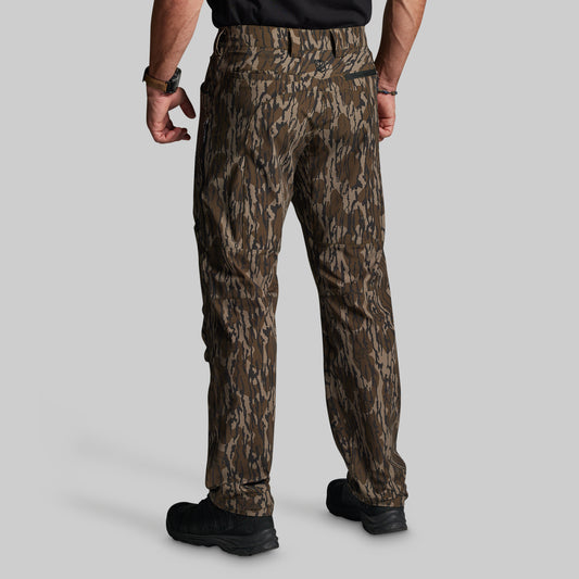 A man shown from the back wearing camouflage outdoor pants, a black shirt, and black boots against a solid gray background.