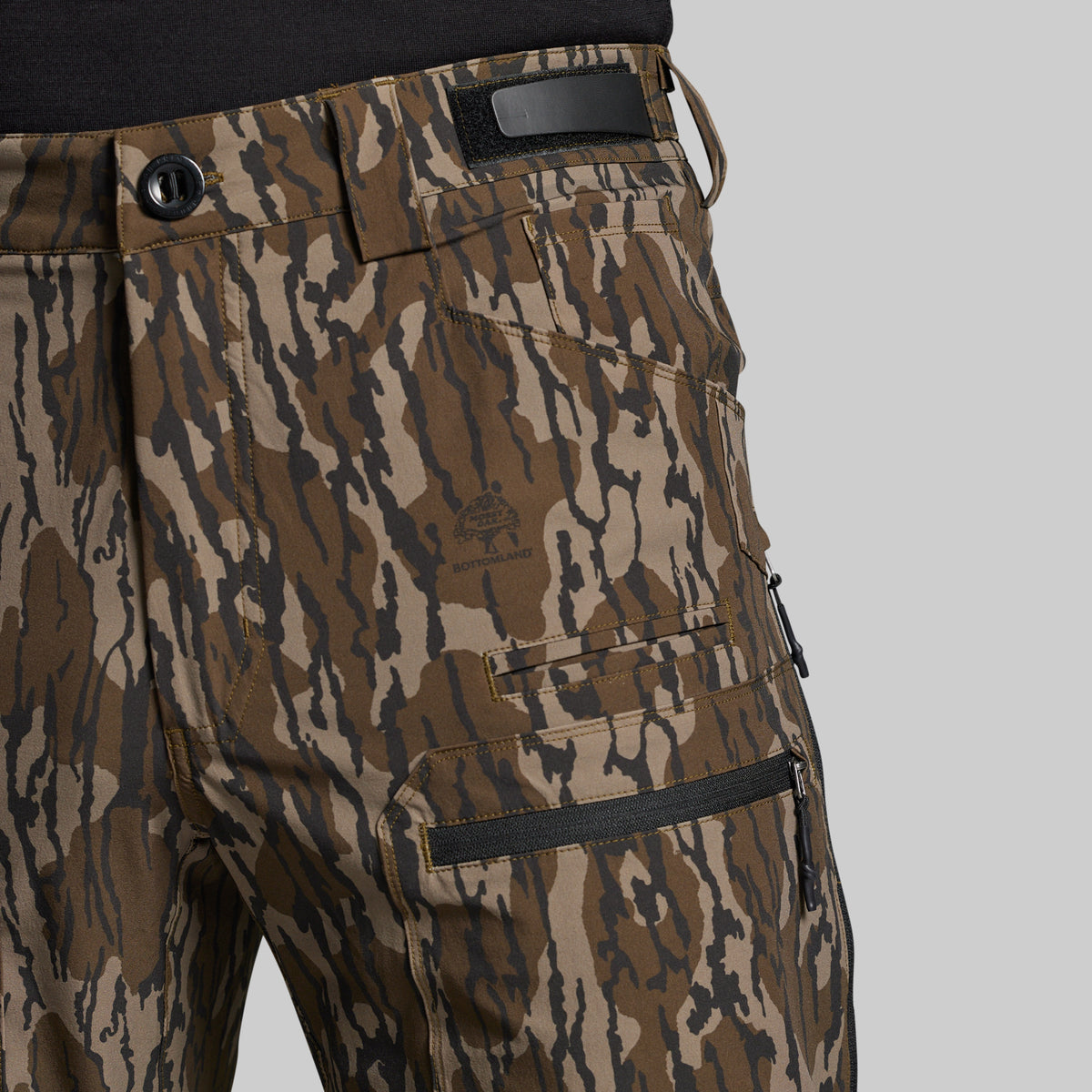 Close-up of a pair of camouflage pants with a brown, tan, and black pattern, featuring a zippered cargo pocket and a black waistband patch, shown on a model wearing a black shirt against a light gray background.