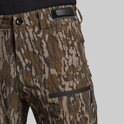 Close-up of a pair of camouflage pants with a brown, tan, and black pattern, featuring a zippered cargo pocket and a black waistband patch, shown on a model wearing a black shirt against a light gray background.