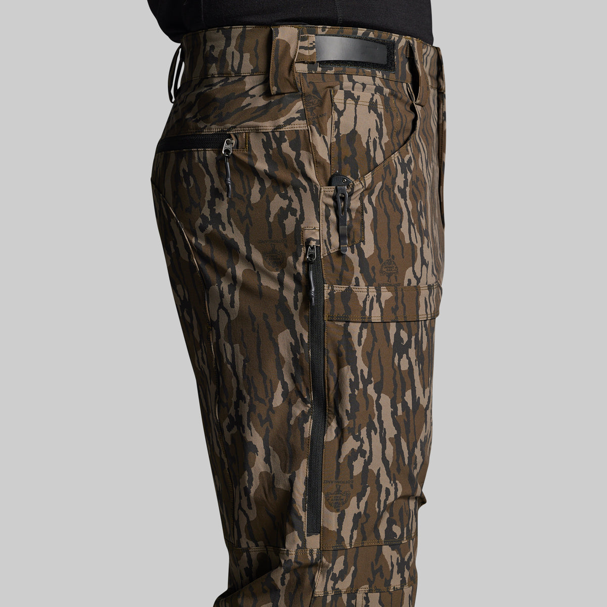 Close-up of a person wearing camouflage cargo pants with multiple zippered pockets and a black belt, shown against a light gray studio background.
