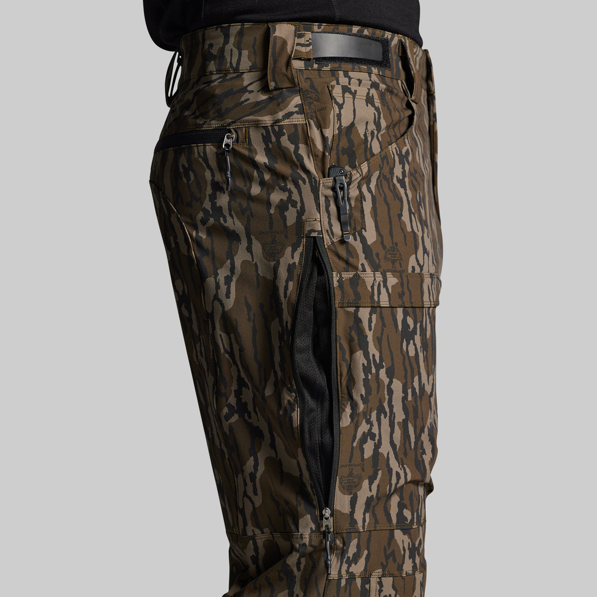 Close-up of a person wearing camouflage cargo pants with multiple zippered pockets and a black belt, shown against a light gray studio background.