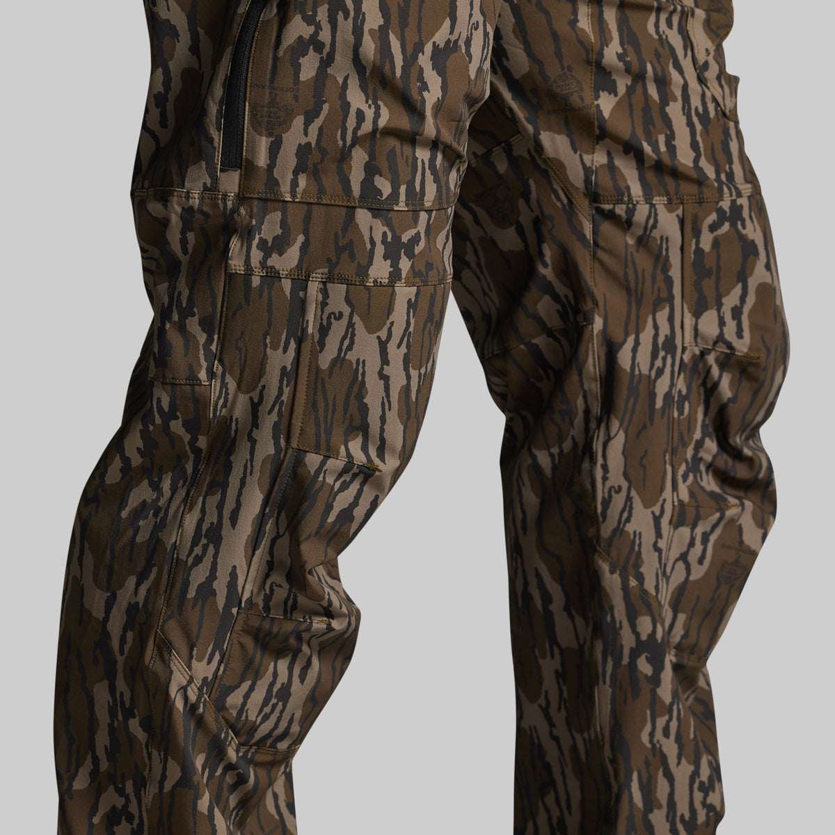 Close-up of a person wearing camouflage pants with a brown, tan, and black bark-style pattern against a light gray studio background.