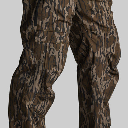 Close-up of a person wearing camouflage pants with a brown, tan, and black bark-style pattern against a light gray studio background.