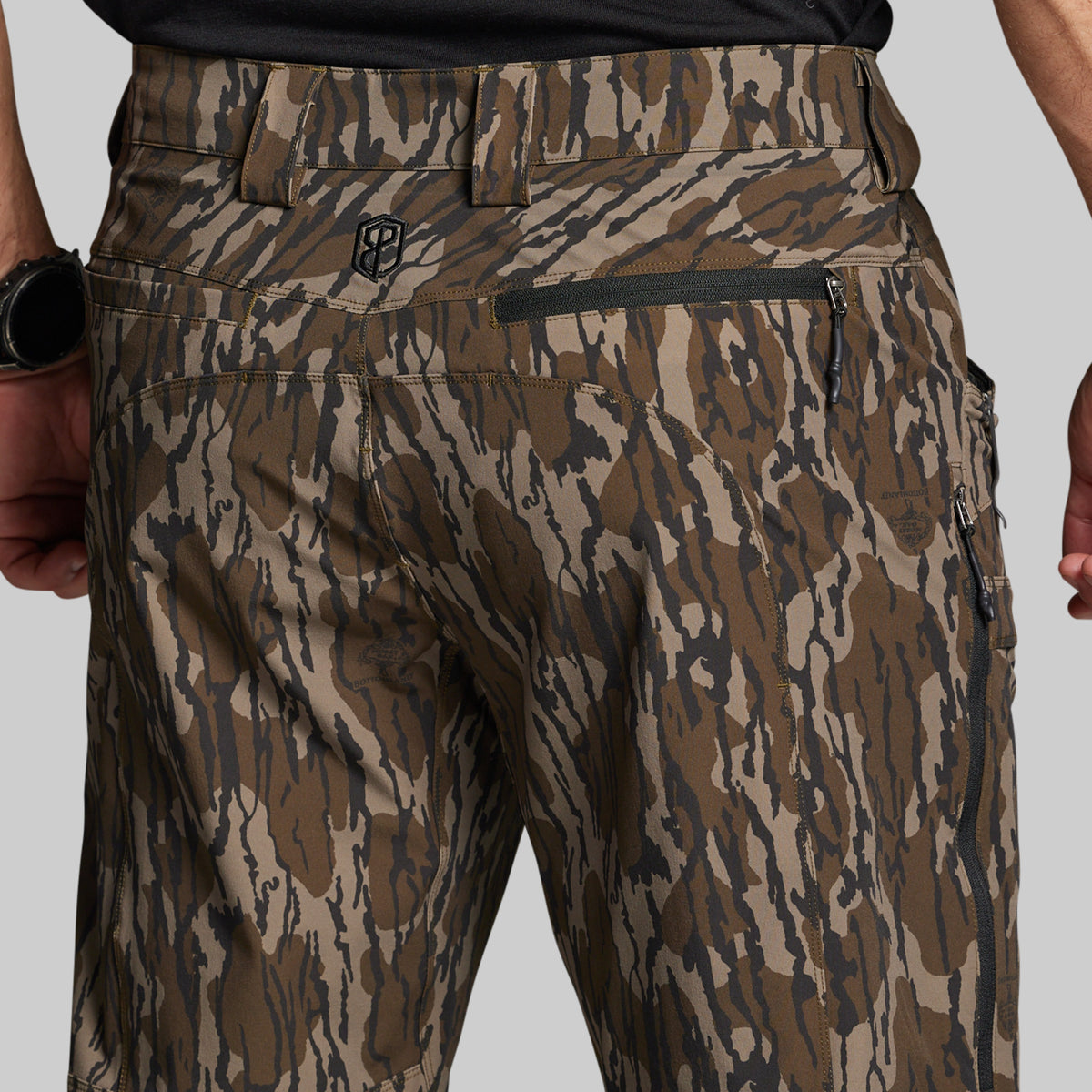 Close-up back view of a person wearing camouflage pants with a brown, tan, and dark gray mossy oak bottomland pattern, shown against a light gray studio background.