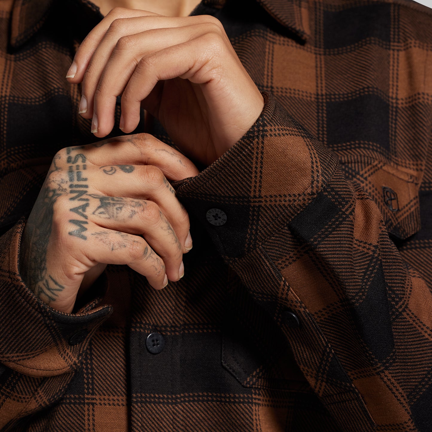 Close-up of a person wearing a brown plaid shirt with visible tattoos on their hands.