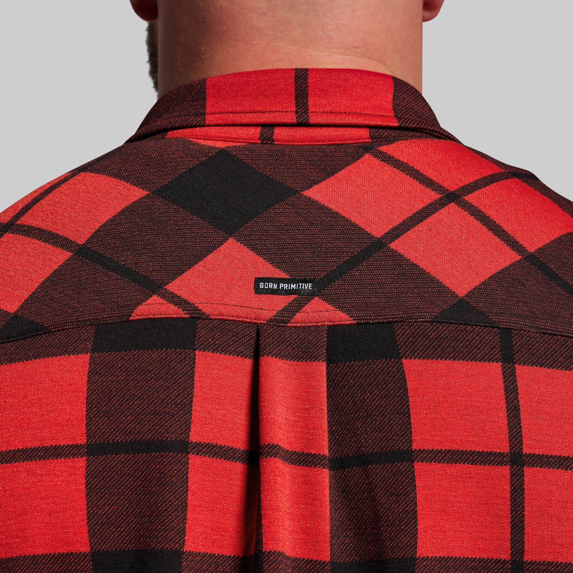 Red and black plaid shirt with 'Born Primitive' logo on a gray background