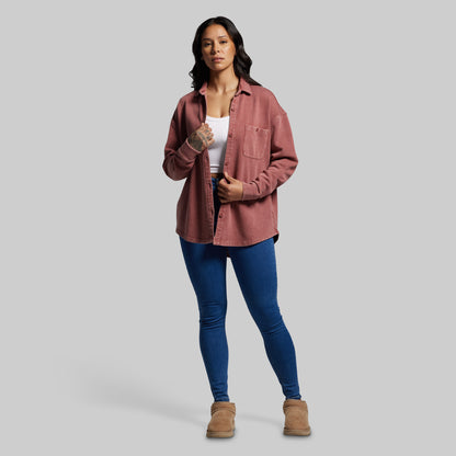 Woman wearing a pink shirt, white top, blue jeans, and brown boots on a gray background