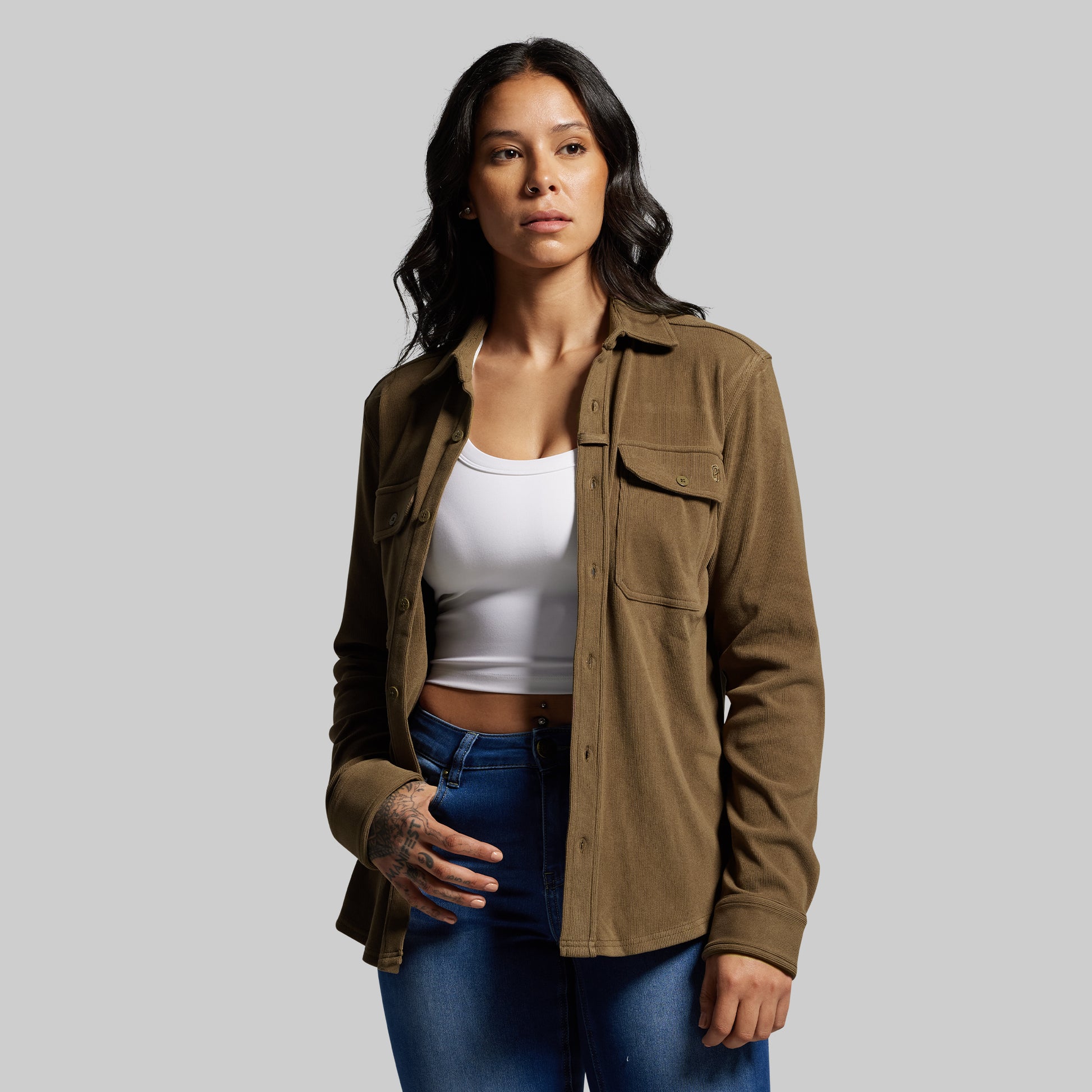 Woman wearing a brown corduroy button-up over a white top and blue jeans on a gray background