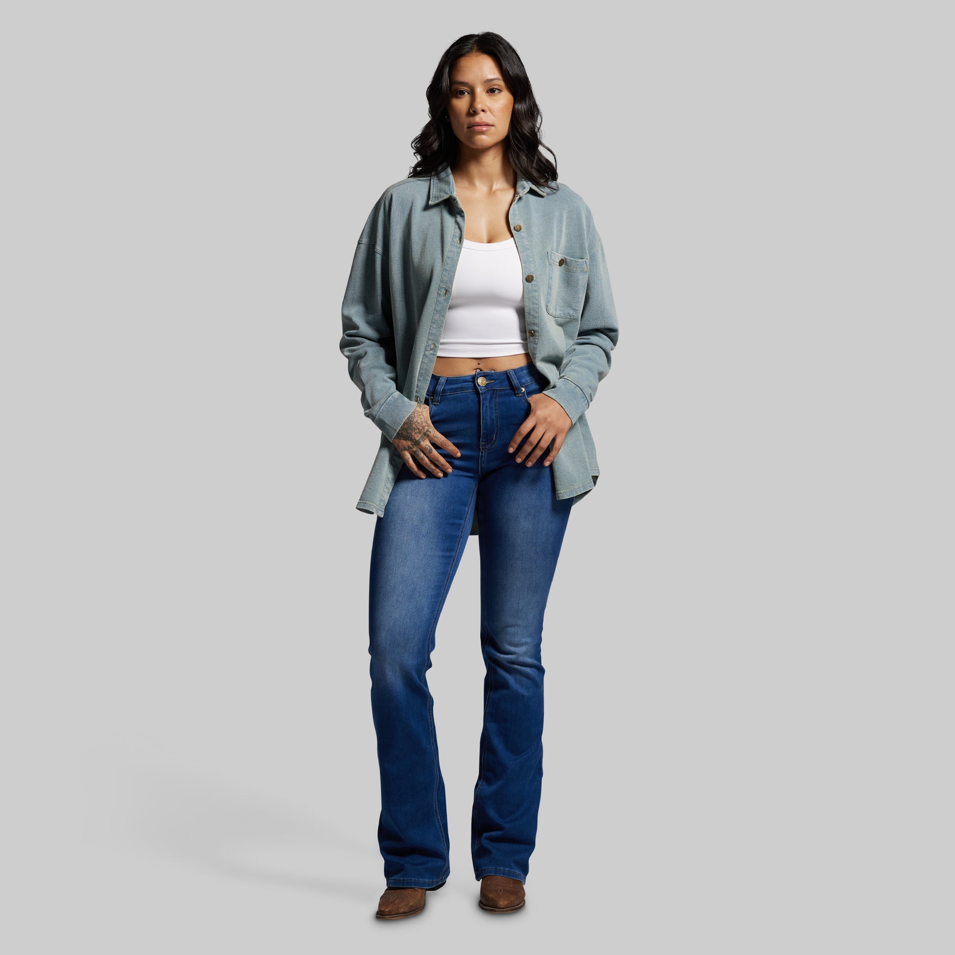 Woman wearing a light blue denim shirt, white top, and blue jeans on a gray background