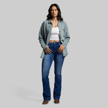 Woman wearing a light blue denim shirt, white top, and blue jeans on a gray background