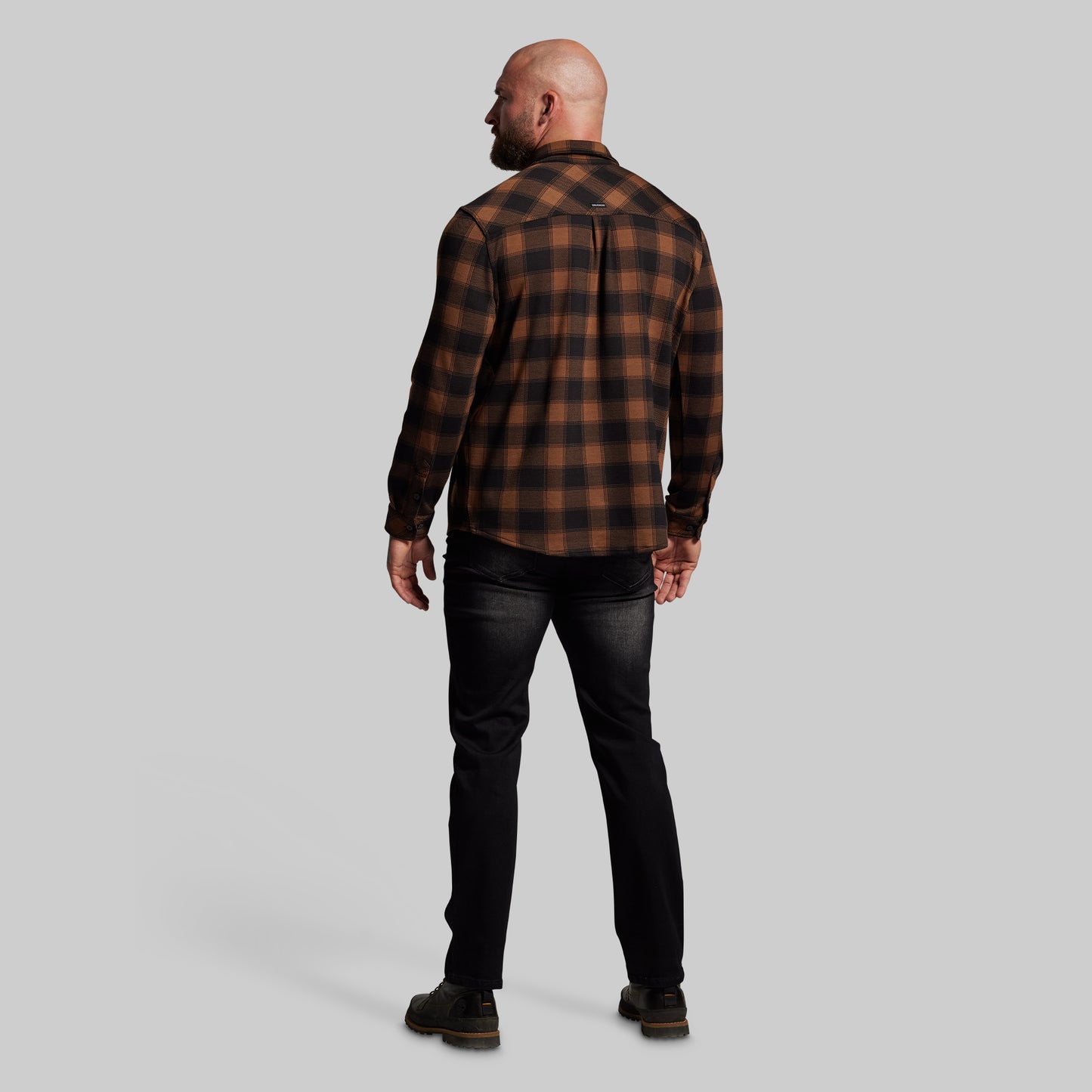 Man wearing a brown and black plaid shirt and black jeans on a gray background