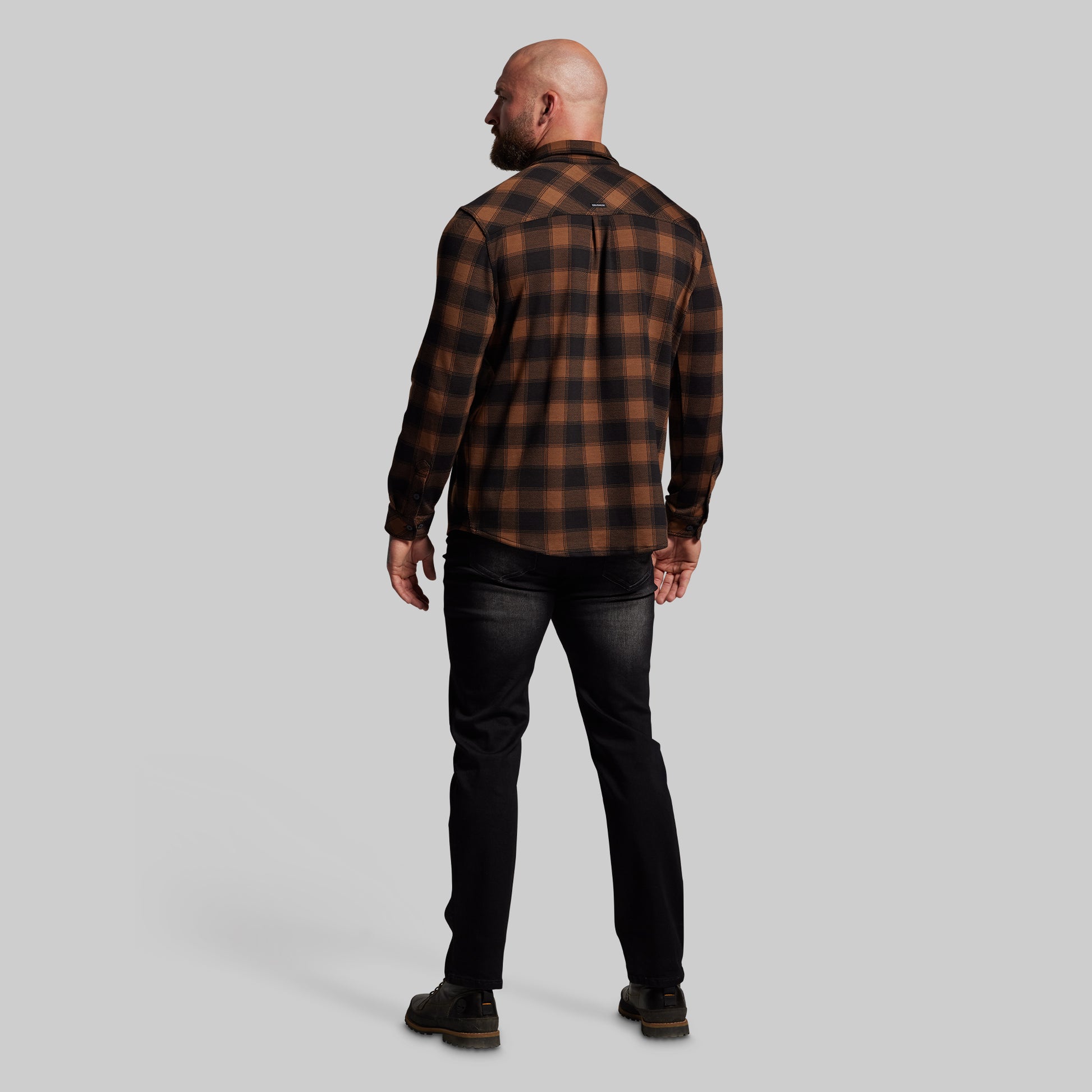 Man wearing a brown and black plaid shirt and black jeans on a gray background
