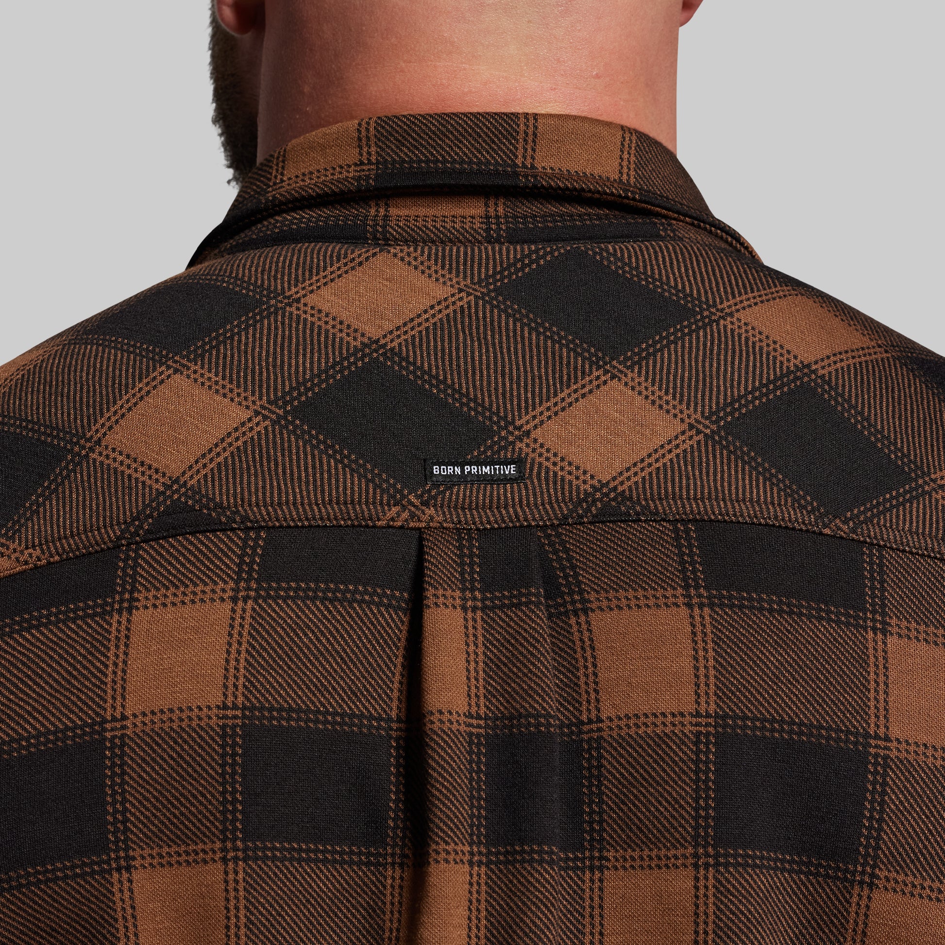 Brown and black plaid shirt with 'Born Primitive' label on a gray background
