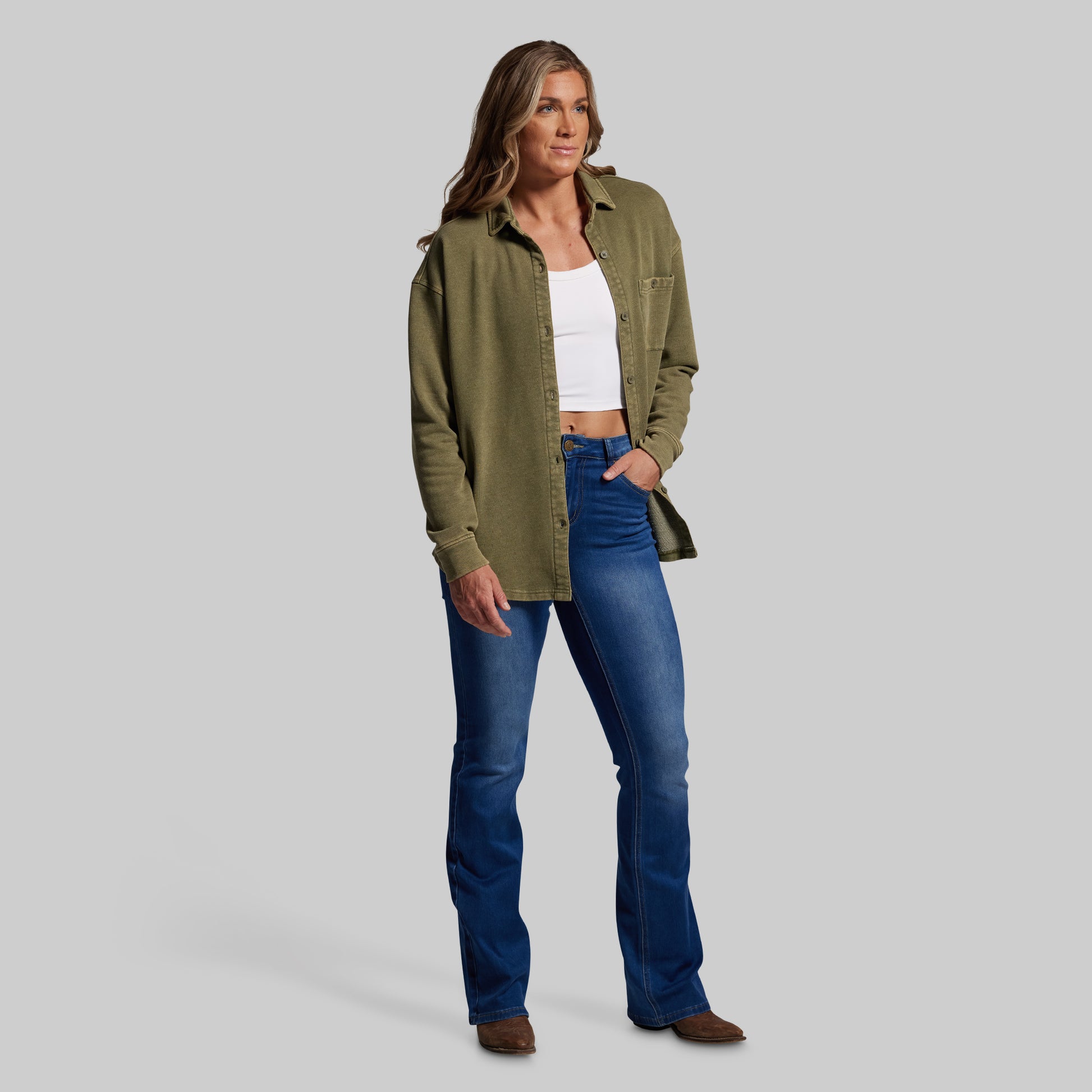 Woman wearing an olive green shirt, white top, and blue jeans on a gray background