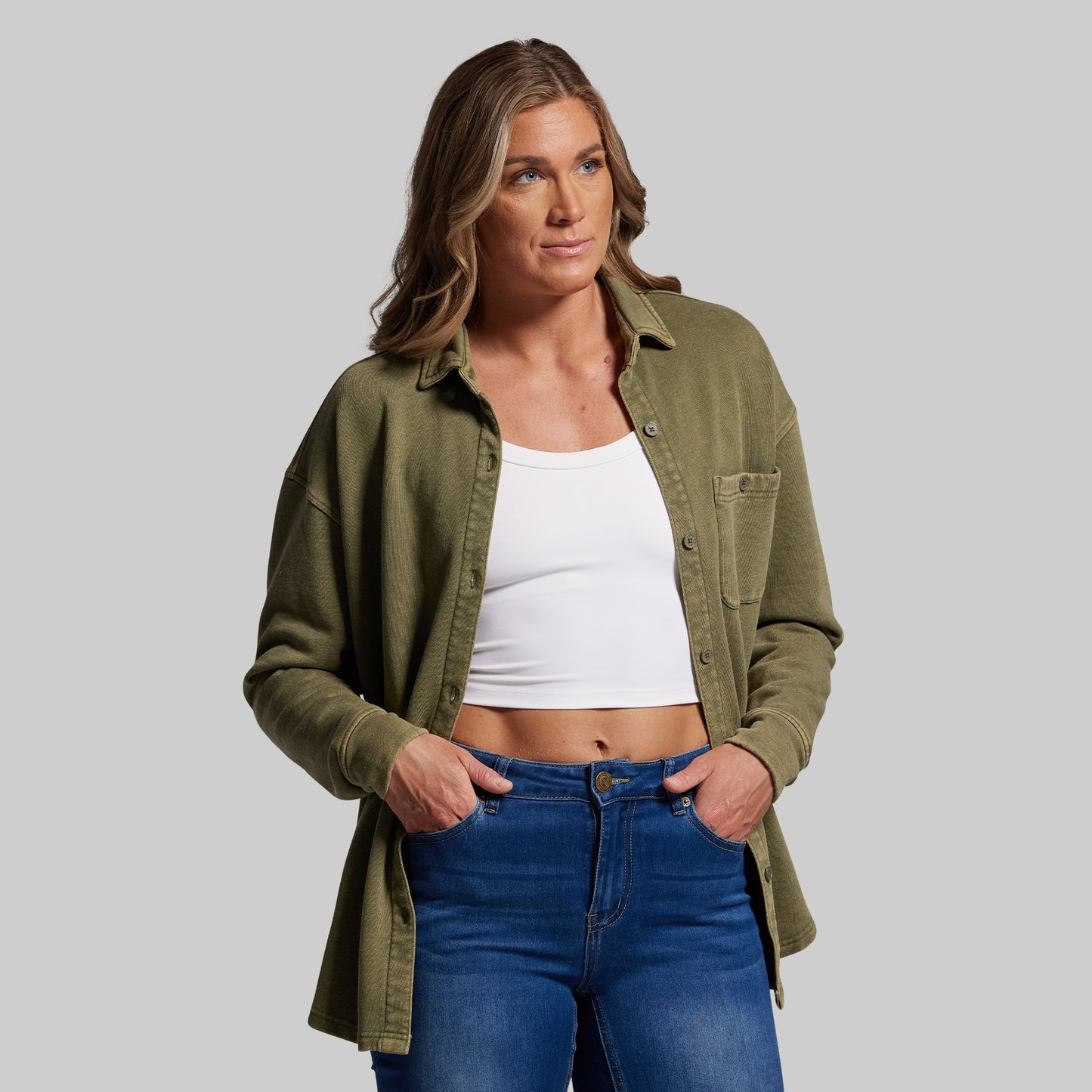 Woman wearing an olive green shirt, white crop top, and blue jeans on a gray background