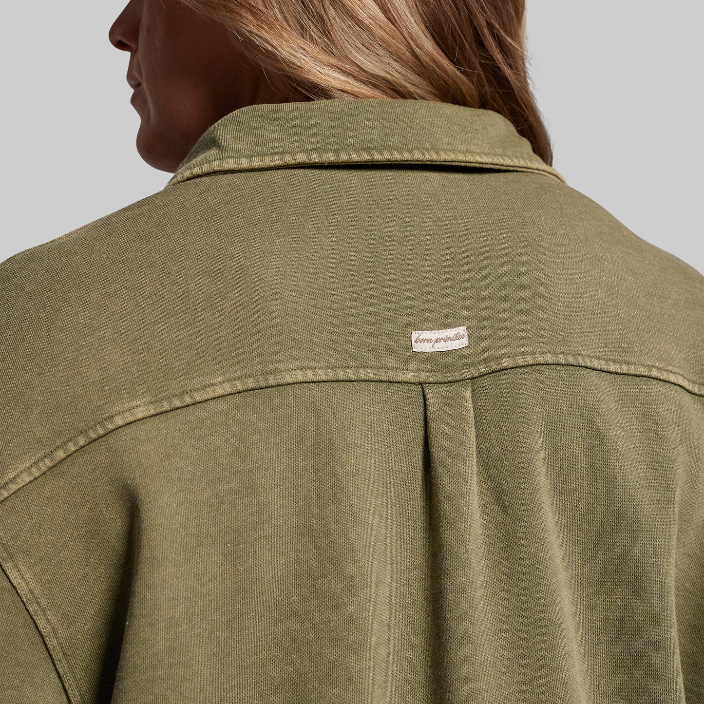 Close-up of a person wearing an olive green shirt with a brand label on a gray background